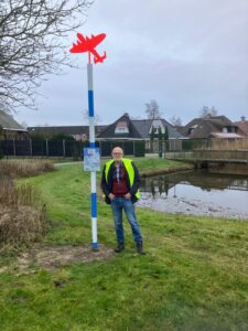 Een man in een geel hesje staat bij een blauw-witgeblokte paal met een rood vliegtuigbordje